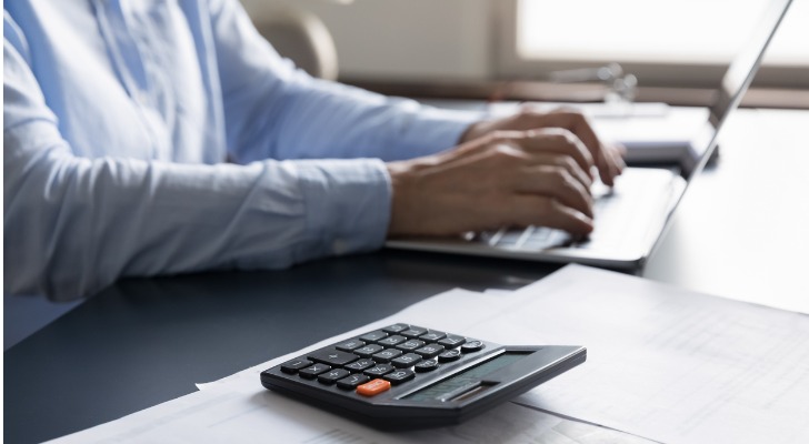 A close-up of a man working on a laptop computer with a calculator in the foreground. 
